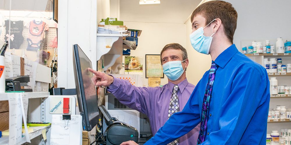 Behind the counter in a pharmacy, two men wearing button-down shirts and slacks stand in front of a computer. They are both wearing blue surgical masks. One man points to information on the computer monitor while the other types on the keyboard.