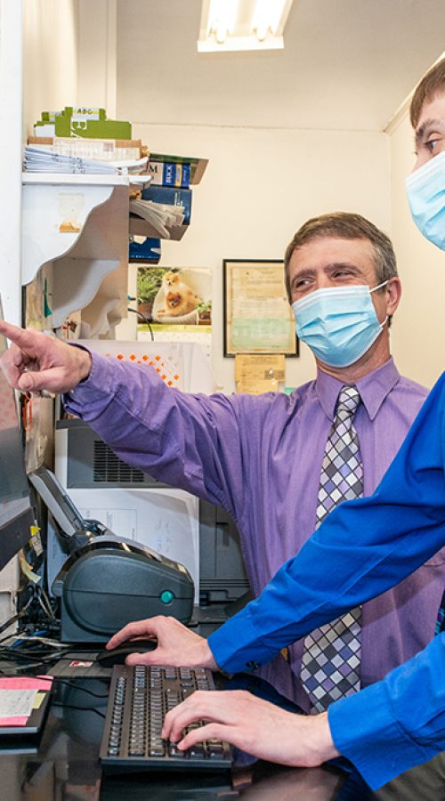 Behind the counter in a pharmacy, two men wearing button-down shirts and slacks stand in front of a computer. They are both wearing blue surgical masks. One man points to information on the computer monitor while the other types on the keyboard.