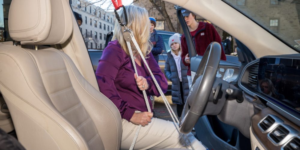 A photo taken from inside, passenger seat of the car looks toward driver seat showing a woman climbing in using a rope pulley. In the background, students observe her use of the tool.