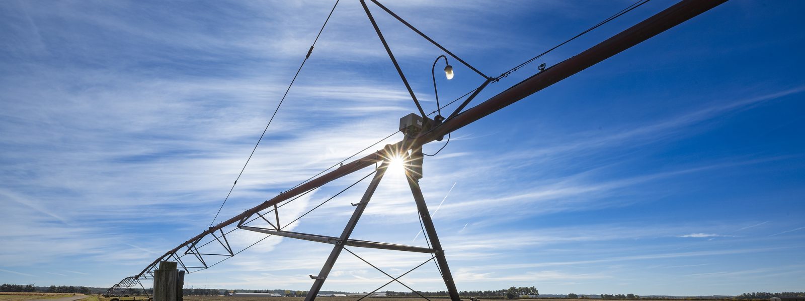 An irrigation pivot sits in a brown potato field under a blue sky.