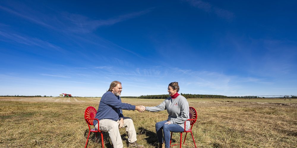 Two people sit in Badger red metal chairs in the middle of a brown potato field under a blue sky. They reach out and shake hands with each other.