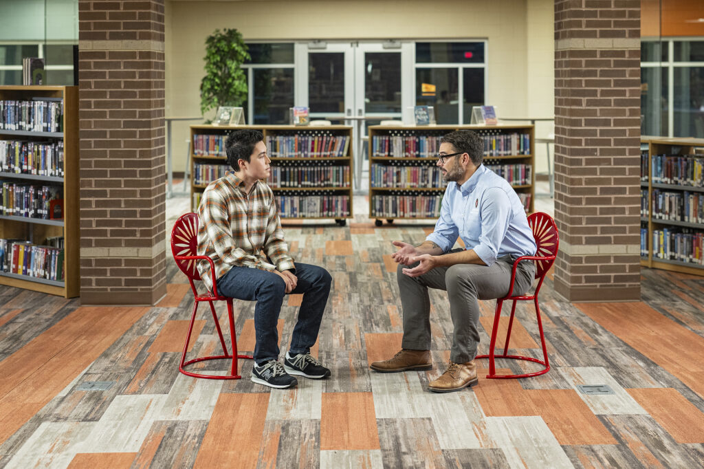 Reed Trueblood (left) and Thomas Owenby (right) talk as they sit in red metal Wisconsin Union chairs in the Dodgeville High School library.
