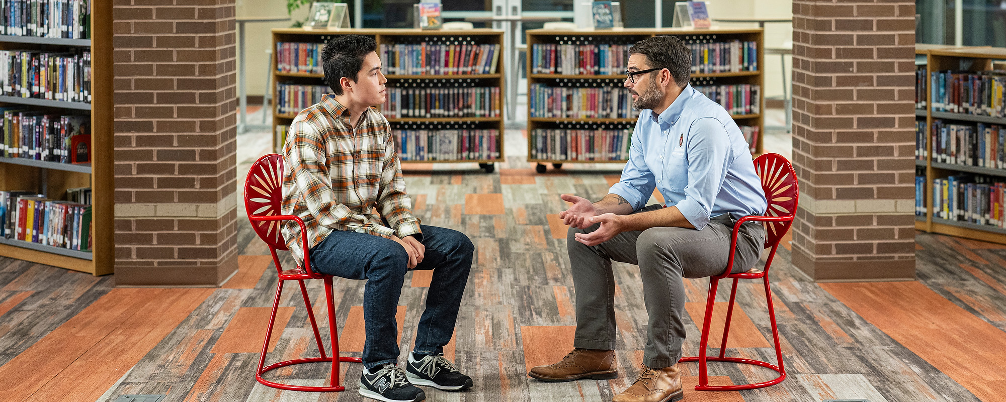 Two people sitting on red Wisconsin Union Sunburst Terrace chairs, talking face-to-face and surrounded by books in a library.