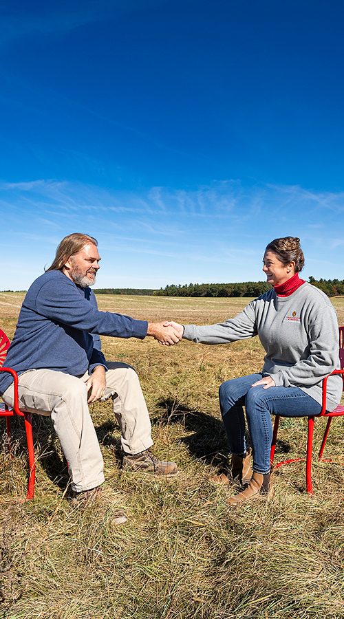 Two people sit in Badger red metal chairs in the middle of a brown potato field under a blue sky. They reach out and shake hands with each other.