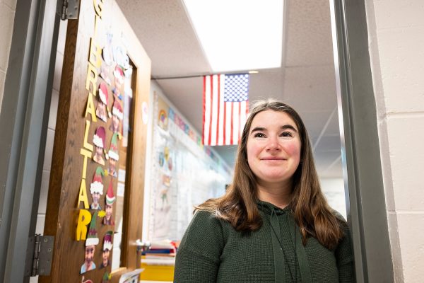 Mackenzie Straub stand in the entrance to her classroom, smiling. The classroom door is decorated with colorful alphabet letters, and an American flag hangs above in the background.
