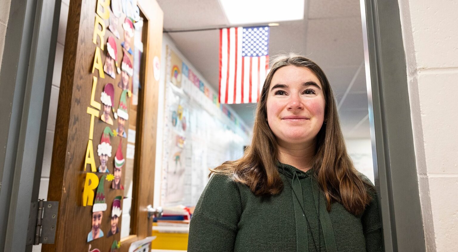 Mackenzie Straub stand in the entrance to her classroom, smiling. The classroom door is decorated with colorful alphabet letters, and an American flag hangs above in the background.