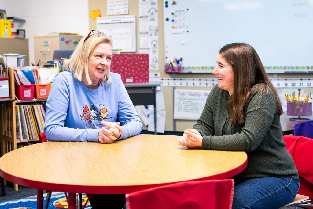 Two individuals are engaged in a conversation at a red table in a colorful classroom setting.