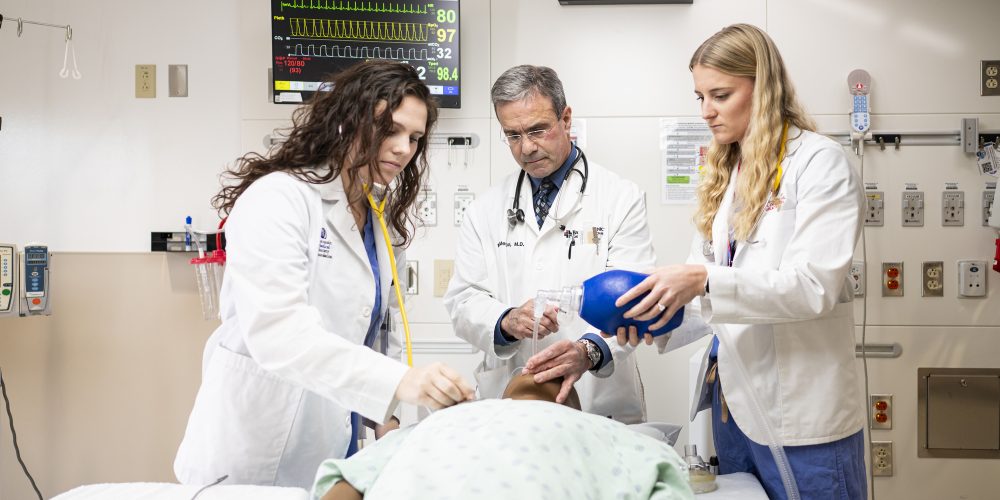 Doctor Michael Medich demonstrates procedure on medical dummy lying on a gurney to two medical students, all three wearing white lab coats