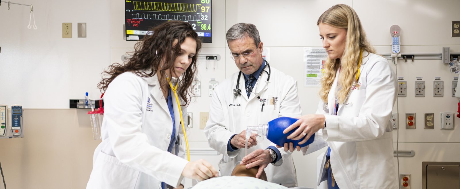 Doctor Michael Medich demonstrates procedure on medical dummy lying on a gurney to two medical students, all three wearing white lab coats