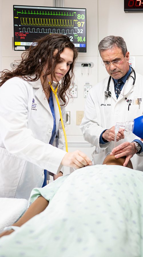 Doctor Michael Medich demonstrates procedure on medical dummy lying on a gurney to two medical students, all three wearing white lab coats