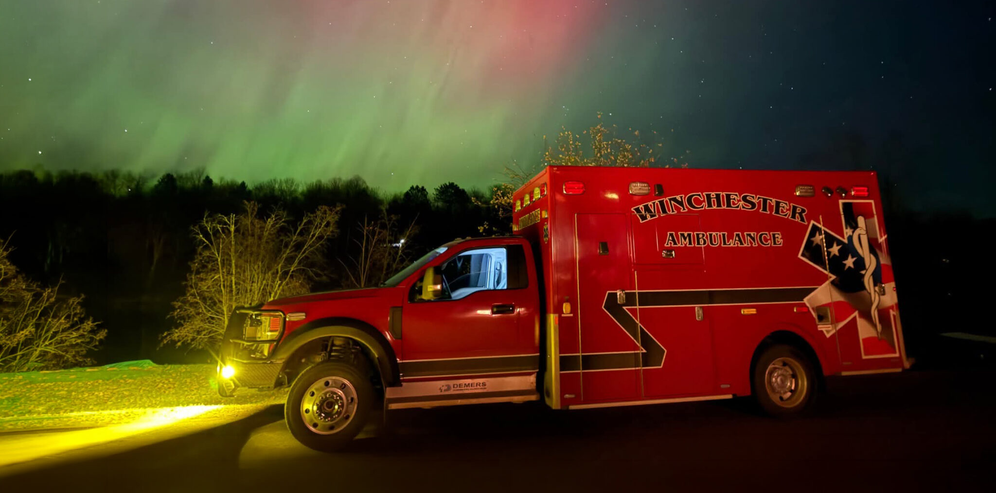 A red ambulance with the words "Winchester Ambulance" on the side of the road at night with Northern Lights in the background