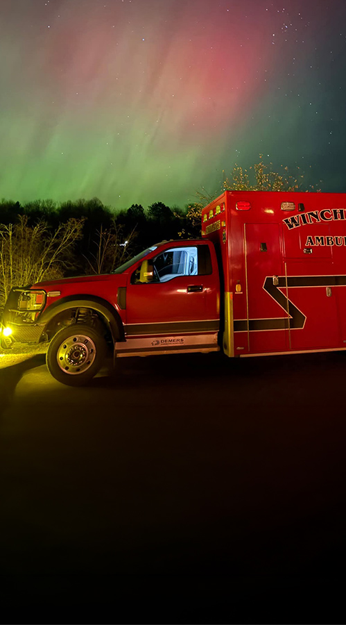 A red ambulance with the words "Winchester Ambulance" on the side of the road at night with Northern Lights in the background
