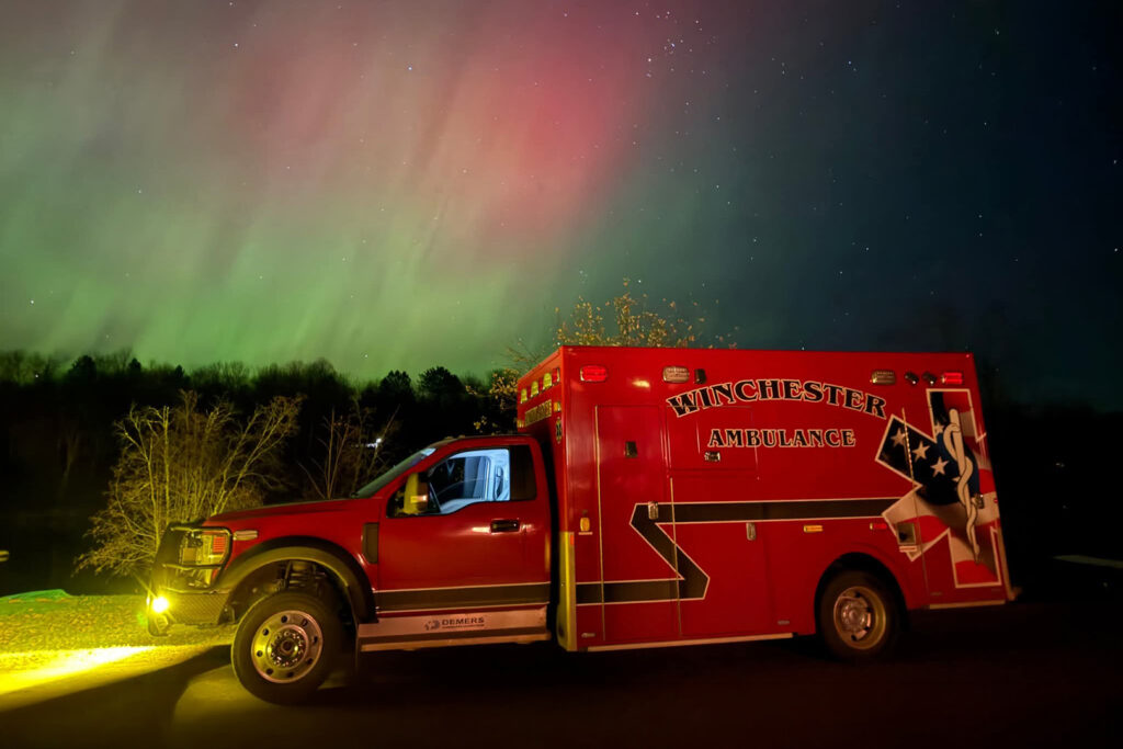 A red ambulance with the words "Winchester Ambulance" on the side of the road at night with Northern Lights in the background