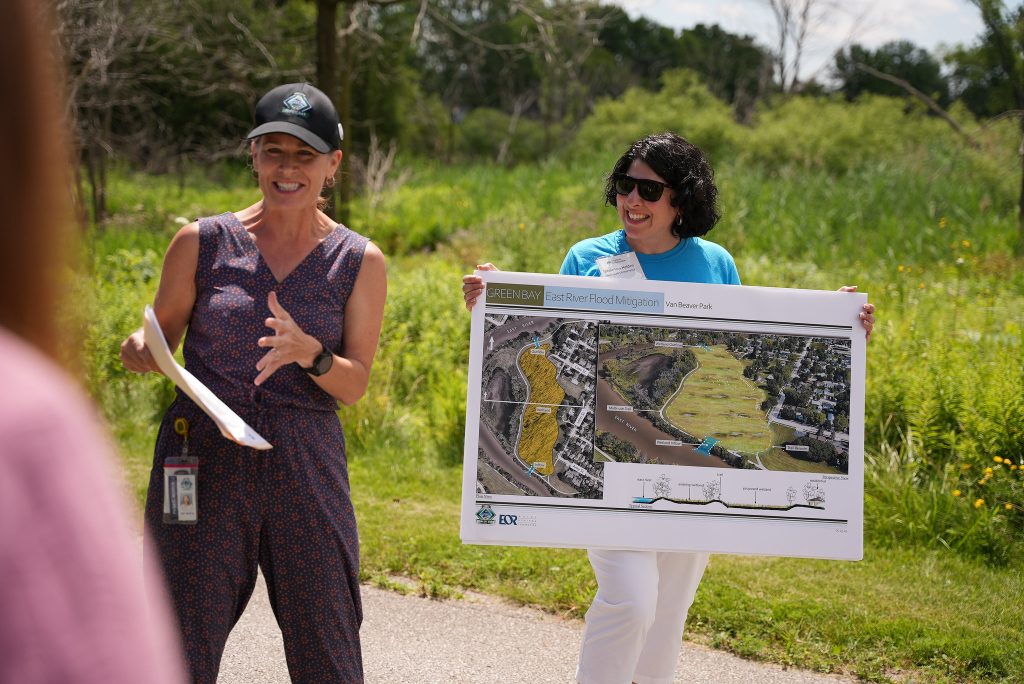 Two people stand along a paved path in a grassy natural area during an outdoor presentation. One person speaks while holding papers, and the other holds a large display board showing maps and design plans labeled “Green Bay East River Flood Mitigation, Van Beaver Park.” Trees and vegetation fill the background on a sunny day.